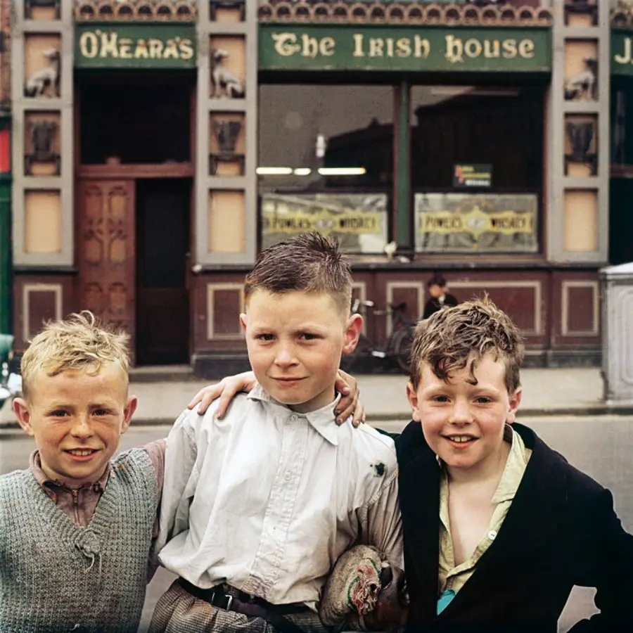 Three smiling boys stand together on a street in front of a pub called “O’Heara’s The Irish House.” The boys have their arms around each other and are dressed in vintage clothing.