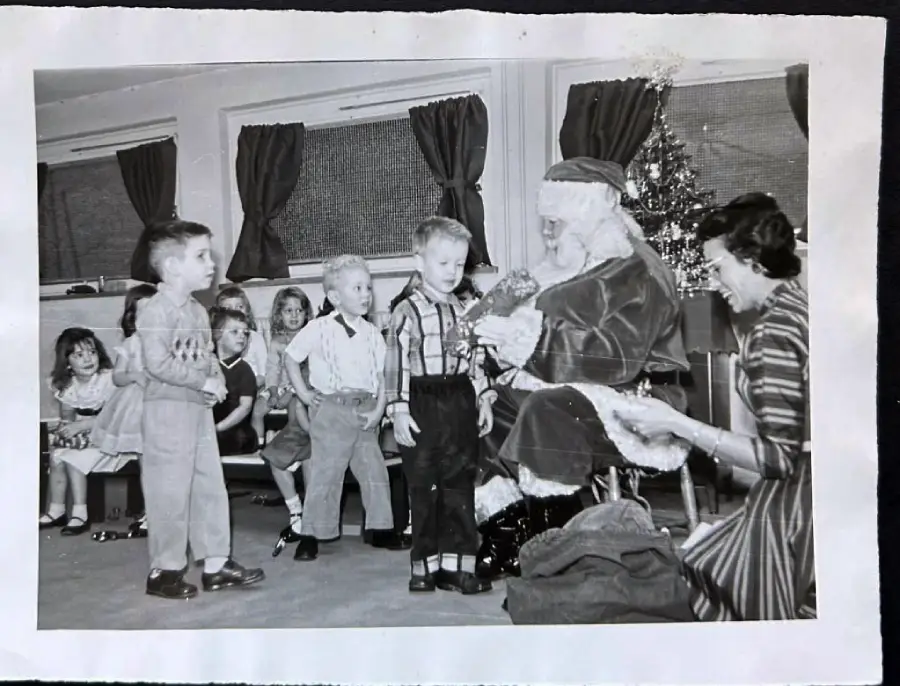 Black-and-white photo of children lined up to meet Santa Claus, who is seated with a woman nearby. Some kids are smiling, others watching. A Christmas tree is in the background.