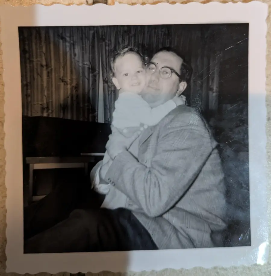 A man wearing glasses holds a smiling baby in his arms. They are indoors, and the photo is in black and white, with a scalloped border.