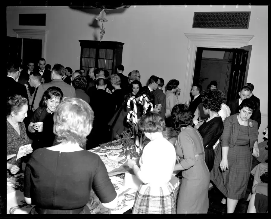 Black and white photo of people socializing at a crowded indoor gathering. Some stand and talk while others gather around a table with food and drinks. The atmosphere is lively and festive.