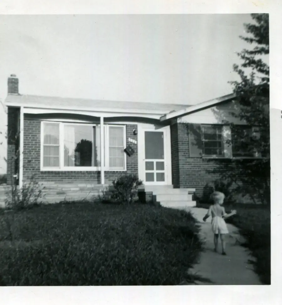 A small child stands on a walkway leading to a brick house with large windows and a front porch. The photo is black and white, and some trees and shrubs are visible in the yard.