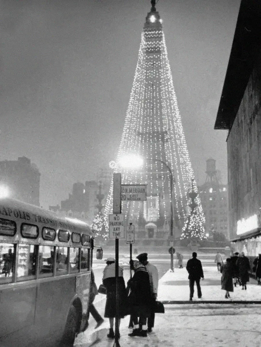 People walk near a city bus and a large, illuminated Christmas tree in a snowy urban square at night; festive lights decorate the scene.