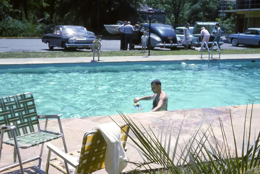 A man swims in a pool near the edge, with lawn chairs and a towel in the foreground. Several vintage cars and people are visible in the parking lot behind the pool.