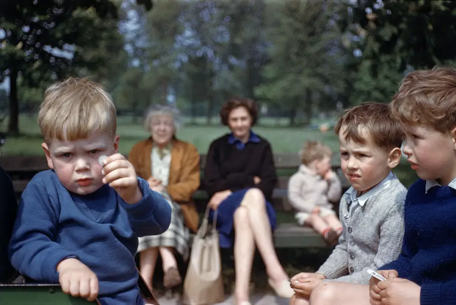 Three young boys sit on a bench in the foreground, one holding an object toward the camera. Behind them, two women and two children sit on another bench in a park, trees and greenery in the background.