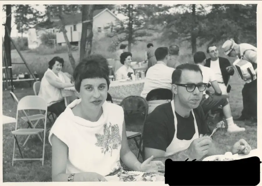 A woman and man sit at a picnic table eating, surrounded by people outdoors. The scene appears to be a casual gathering in a backyard with folding chairs and tables. The photo is in black and white.