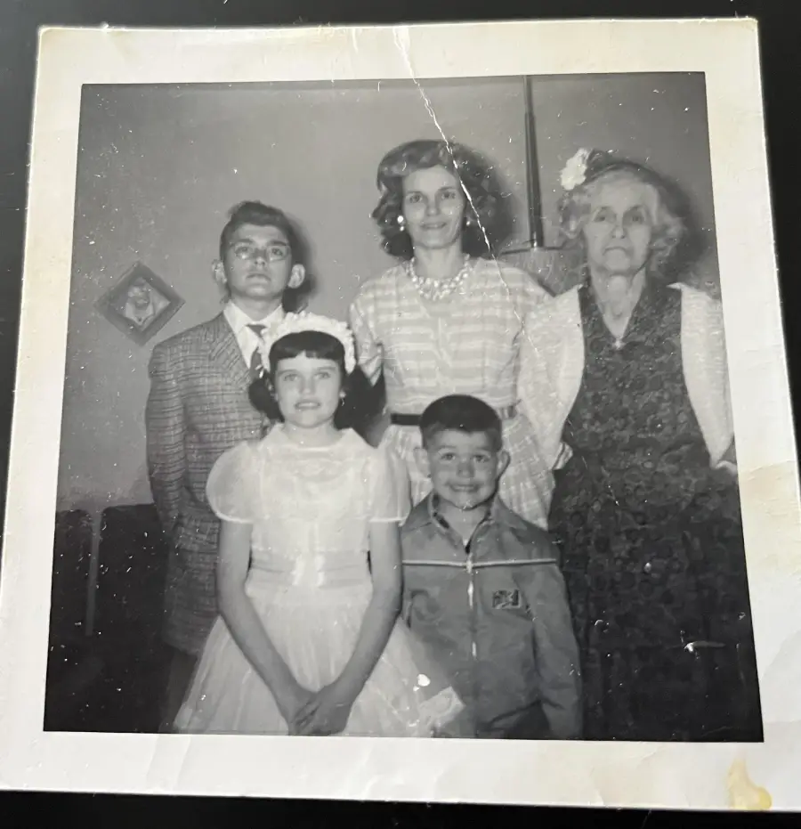A black-and-white photo shows three women (one elderly) and two children. The group is dressed formally and stands indoors, posing for the camera. The photo has some visible creases and wear.