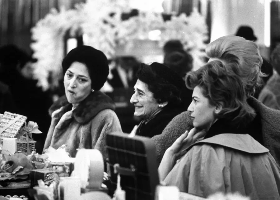 Three women in elegant coats and hats are gathered at a crowded cosmetics counter, surrounded by boxes and mirrors. They appear engaged, with two smiling and one looking thoughtful. The background is softly lit and decorated.