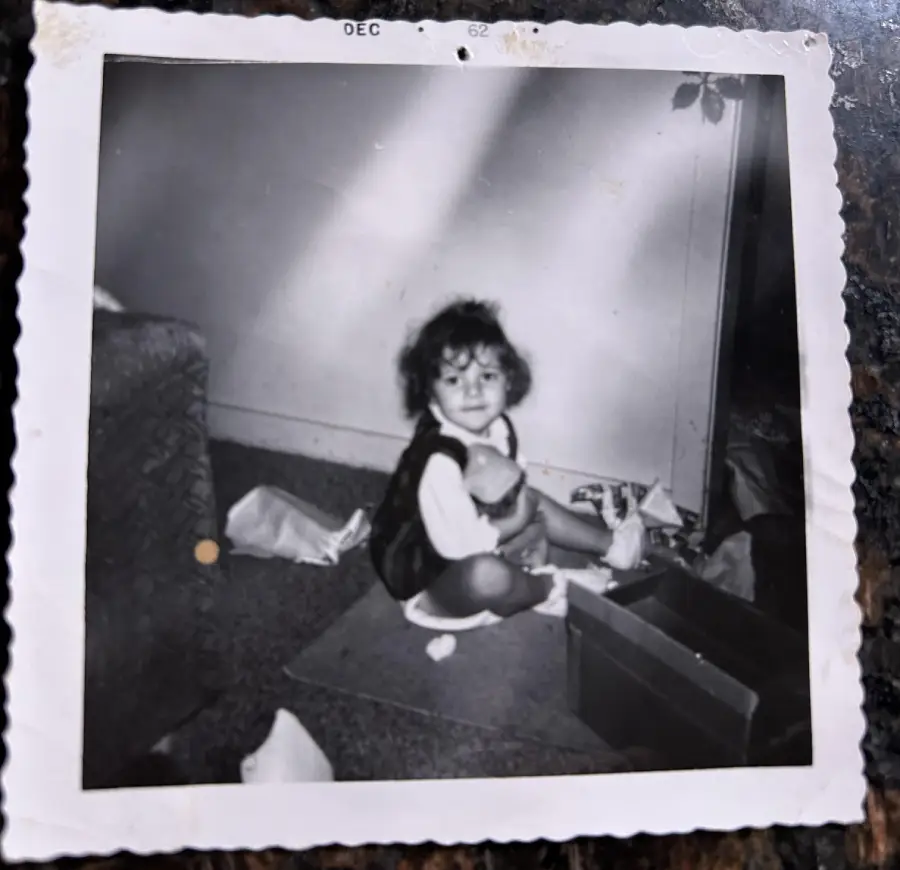 Black-and-white photo of a young child sitting on the floor, holding a doll, surrounded by toys and papers. The image has a white border with a scalloped edge and a date stamp from December 1962 at the top.
