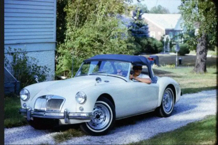 A person wearing sunglasses sits in a white vintage convertible car parked on a gravel driveway, with green trees and houses visible in the background.