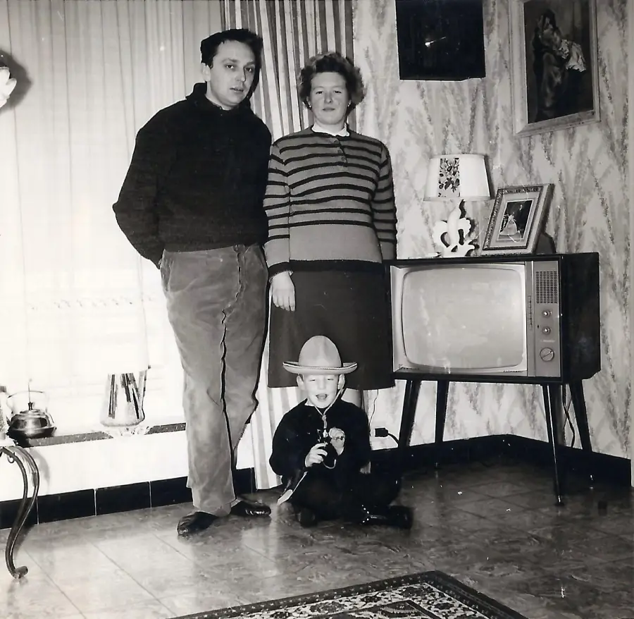 A man and woman stand beside an old TV in a vintage living room. A young boy in a cowboy hat sits on the floor, smiling. The room has patterned wallpaper, framed photos, and a small rug.