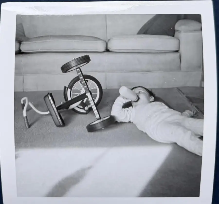 A baby in pajamas lies on a carpeted floor drinking from a bottle beside a toppled tricycle, with a sofa in the background. The photo is black and white.