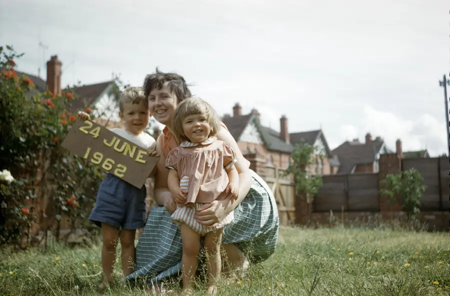 A woman kneels in a grassy yard, hugging two young children. One child holds a sign reading “24 June 1962.” Brick houses and a fence are visible in the background.