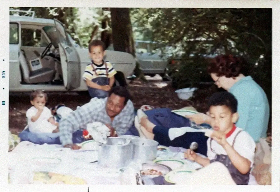 A family enjoys a picnic on a blanket outdoors, with food and pots spread out. Two children sit, one child stands, and two adults relax nearby. An open car door is visible in the background under trees.