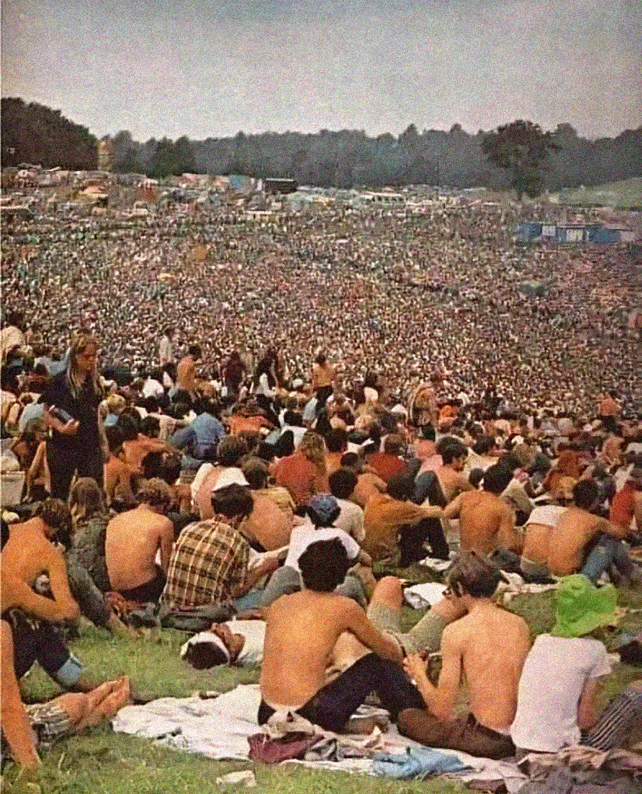 A large crowd sits and lounges on a grassy hill at an outdoor music festival, many people are shirtless and appear relaxed, with tents and trees visible in the background under a cloudy sky.
