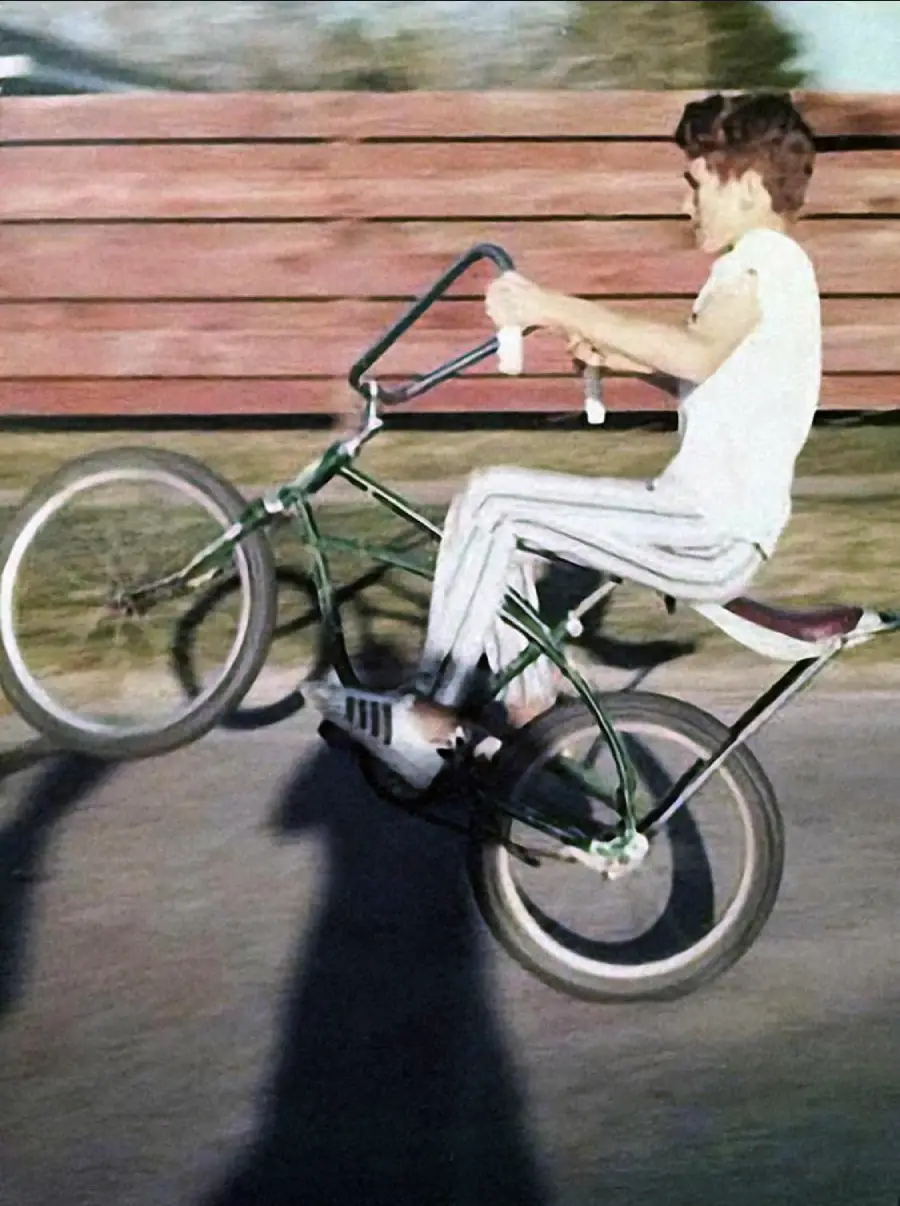 A boy in a white T-shirt and striped pants does a wheelie on a green bike, riding on a street with a wooden fence and trees in the background.