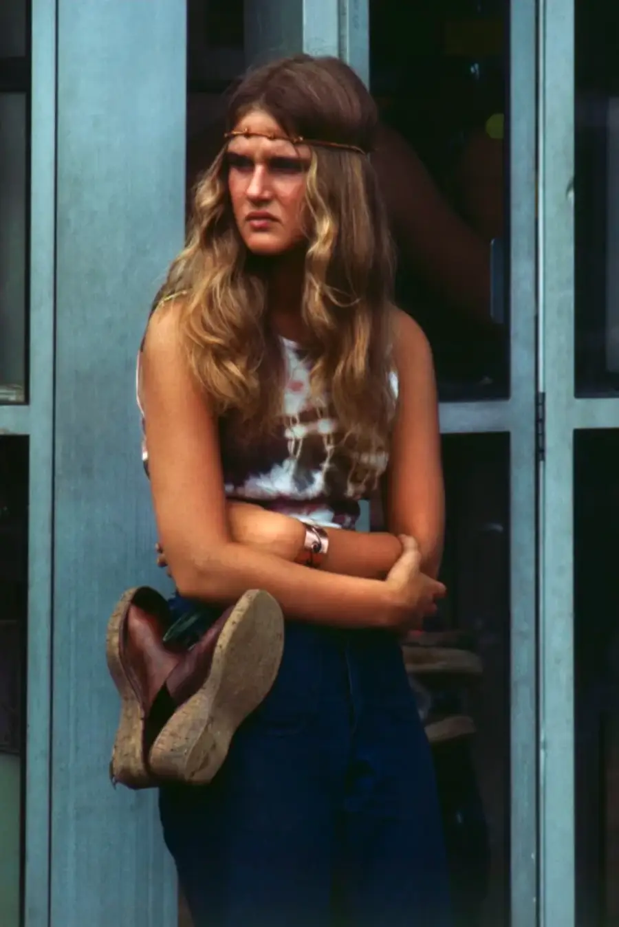 Young woman with long wavy hair and a headband stands with folded arms, holding sandals behind her back, wearing a tie-dye shirt and jeans, in front of glass doors.