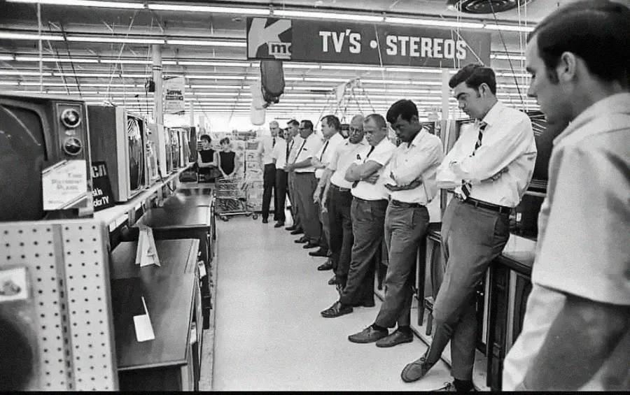 A group of men in shirts and slacks stand in a row inside a store, watching TV sets under a sign that reads “TV’s - STEREOS.” The store appears brightly lit and filled with electronics.