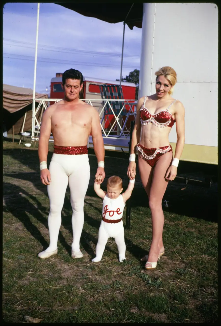 A man, woman, and baby in red and white circus costumes stand on grass, holding hands, with tents and a trailer in the background. The baby’s outfit has “Joe” written on it.