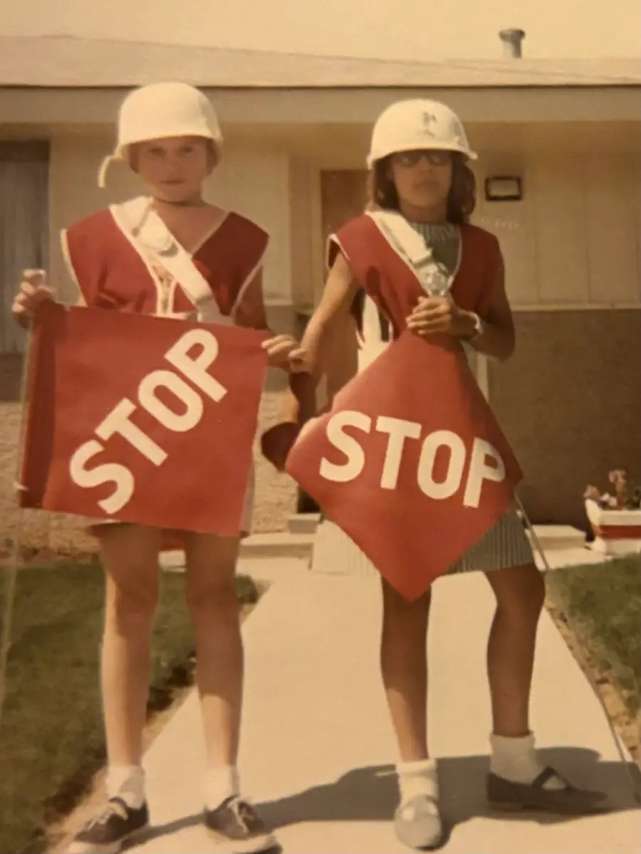 Two children wearing helmets and red sashes hold large “STOP” signs on a sidewalk in front of a house. The photo has a vintage look.
