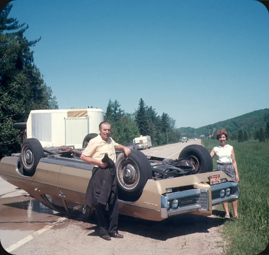 A man and woman stand by an overturned car on a rural road. The car is on its roof next to the grass, with a trailer visible in the background. Both appear unharmed. Trees and hills line the road.
