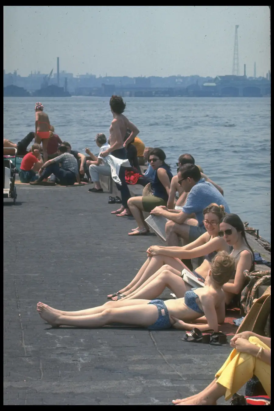 People relax and sunbathe on a pier by the water on a sunny day. Some are sitting, others are lying down, and the city skyline is visible in the background under a hazy sky.