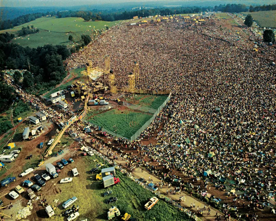 Aerial view of a massive outdoor music festival with a sea of people surrounding a stage in a rural, grassy area, cars and tents scattered around the perimeter.