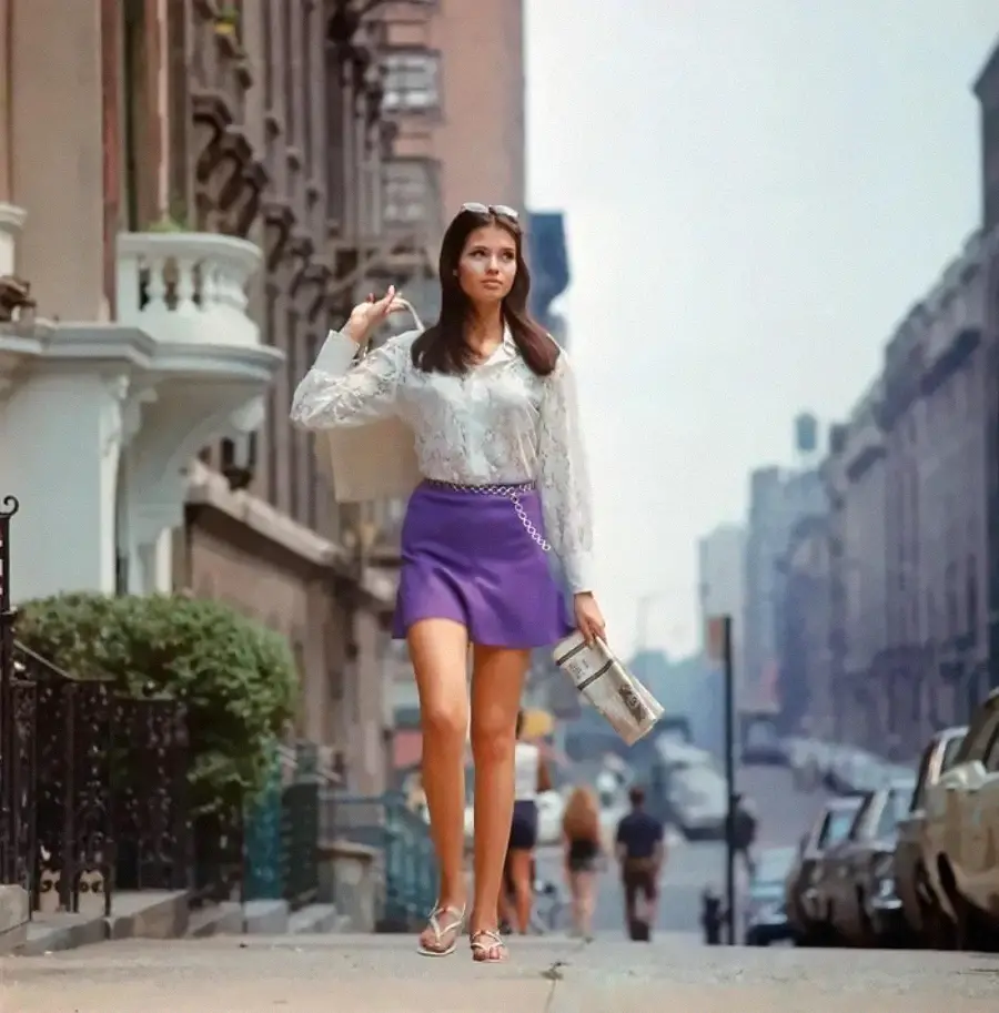 A woman in a white blouse and purple skirt walks on a city sidewalk carrying a bag and a newspaper, with cars and buildings in the background.