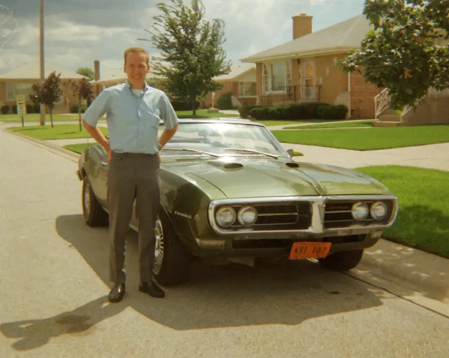 A smiling man stands in front of a green vintage car parked on a suburban street, with houses, lawns, and trees visible in the background.