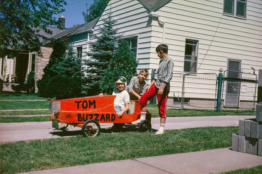 Three children play outside with a homemade red soapbox car labeled “Tom the Buzzard” on a suburban sidewalk, with houses, trees, and a lawn visible in the background.