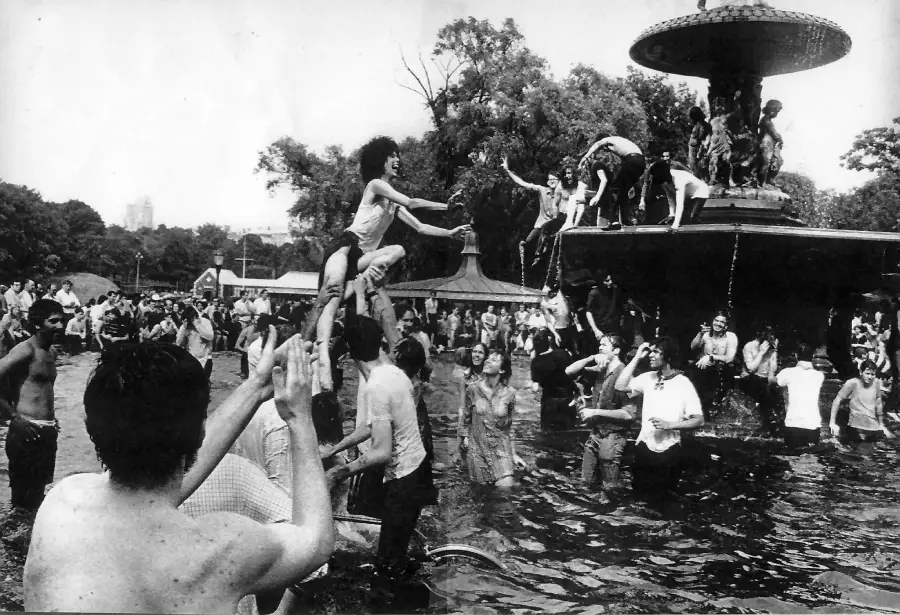A large group of people play and splash in a fountain in a park. Some climb on the fountain, others cheer, while trees and parkgoers fill the background. The scene is lively and energetic.