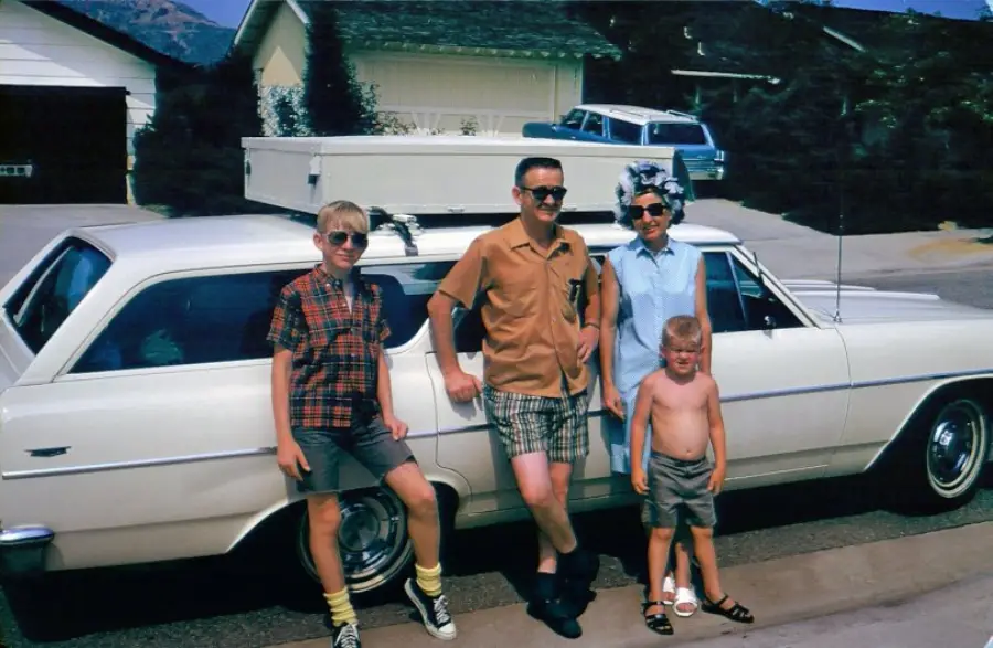 A family of four poses beside a white vintage station wagon in a suburban driveway. The dad and two boys wear summer clothes; the mom is in a dress and sunglasses. A house and car are in the background.