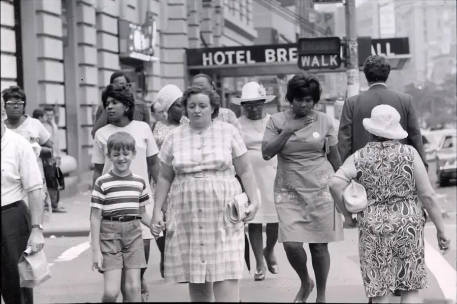 A group of people, including women, a man, and a boy, walk along a city street. Buildings, a “HOTEL BRE” sign, and a “WALK” signal are visible in the background.