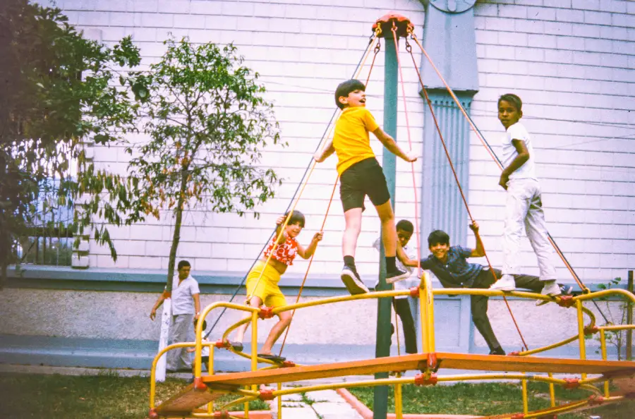 Five children play on a colorful merry-go-round outdoors, with some climbing and others standing. Trees and adults are visible in the background near a white building.