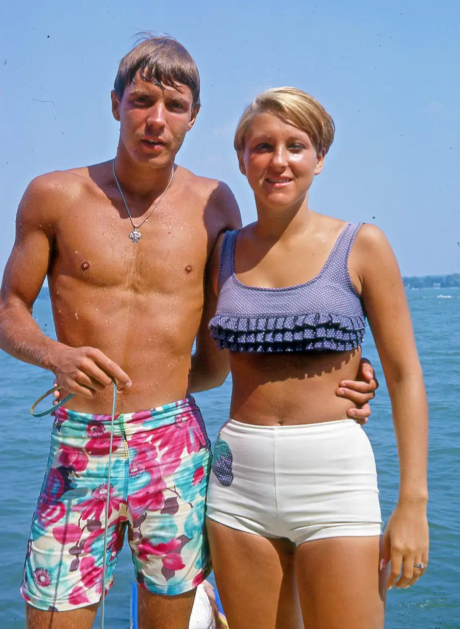 A man and woman in swimsuits stand together by the water on a sunny day, smiling at the camera. The man wears floral swim trunks, and the woman wears a blue crop top with white shorts.