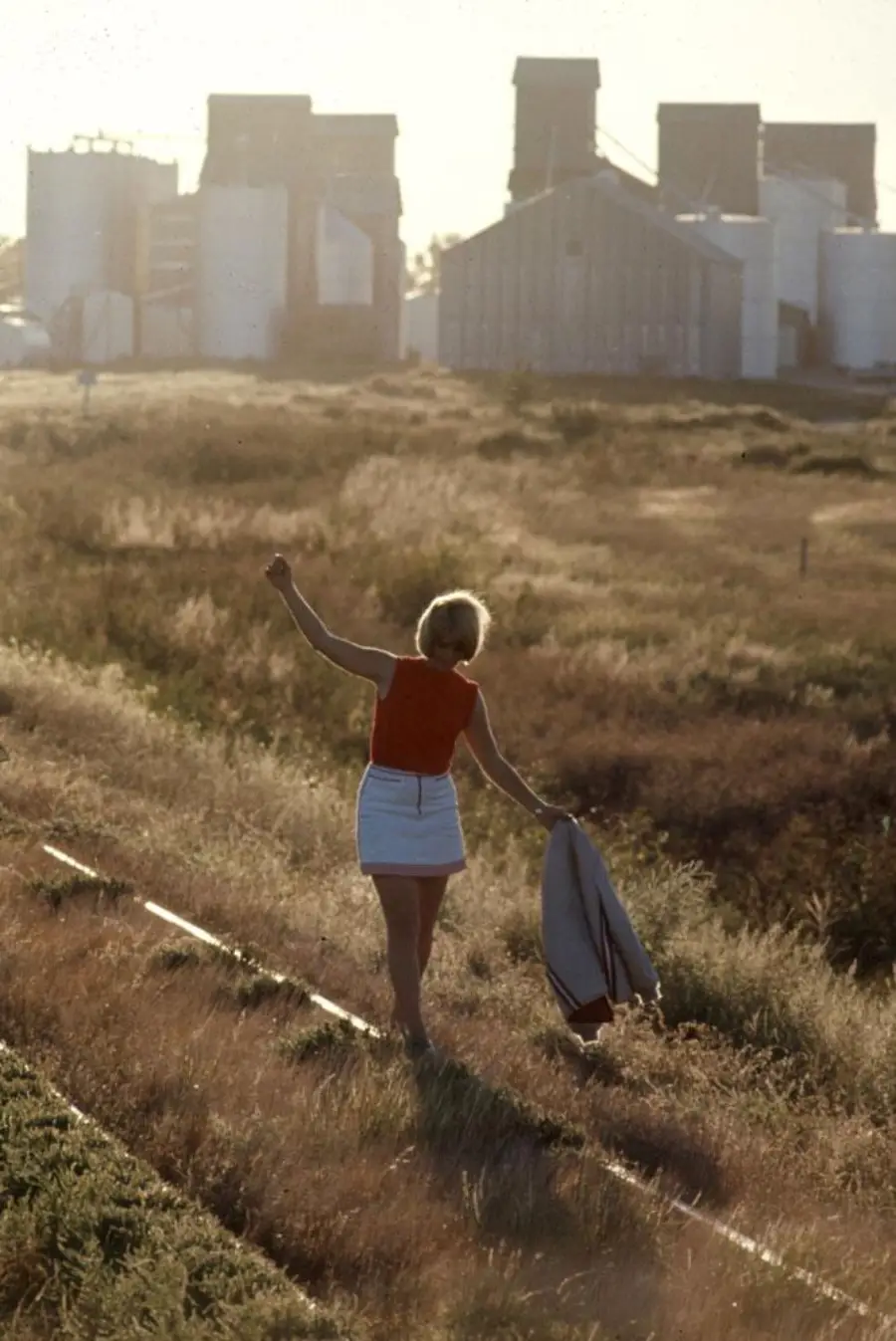 A person in a red top and white skirt walks on railroad tracks through tall grass, holding a jacket, with large grain elevators and silos in the background.