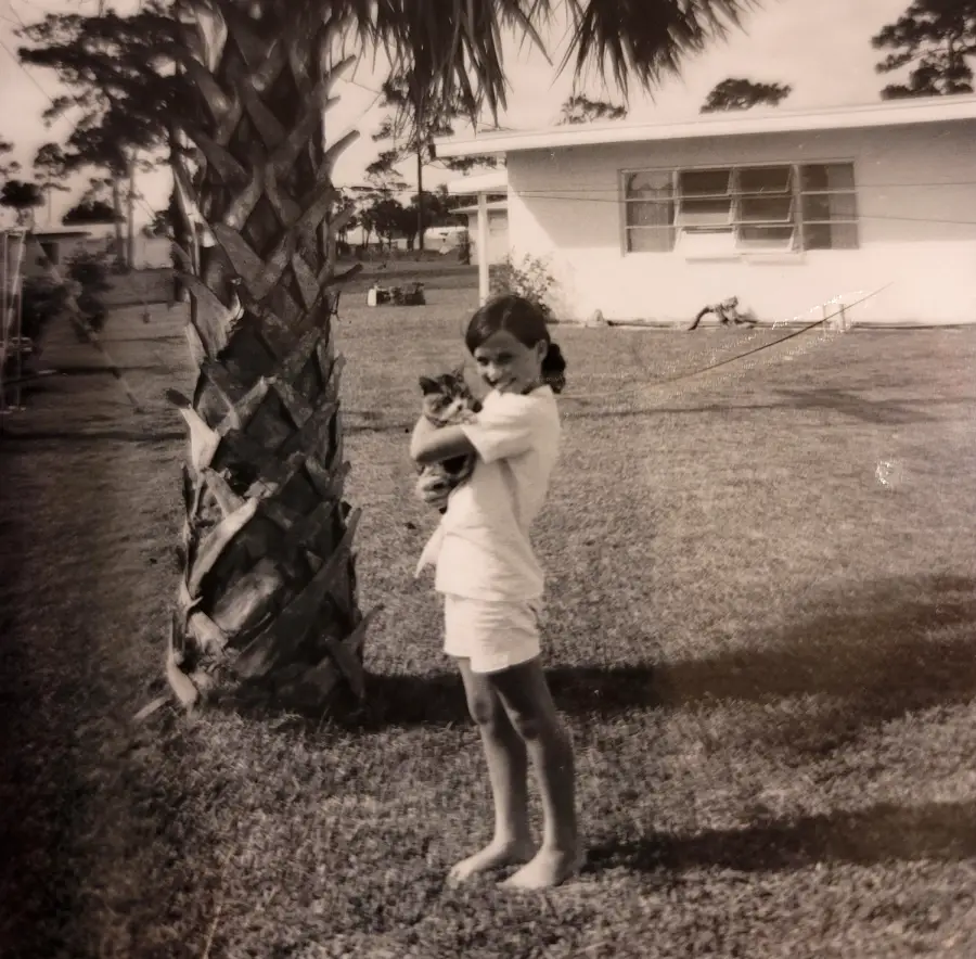 A young girl stands barefoot on grass, holding a cat and smiling. She is beside a palm tree, with a single-story house and more trees visible in the background. The image is black and white.