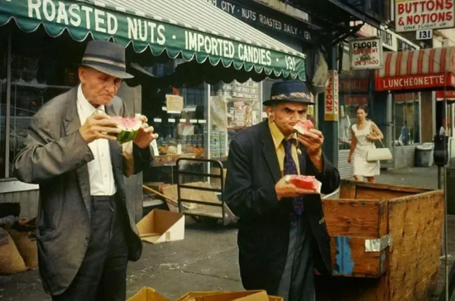 Two older men in suits eat watermelon on a city sidewalk near shops with awnings that read “ROASTED NUTS” and “IMPORTED CANDIES.” Cardboard boxes and a passerby are visible in the background.