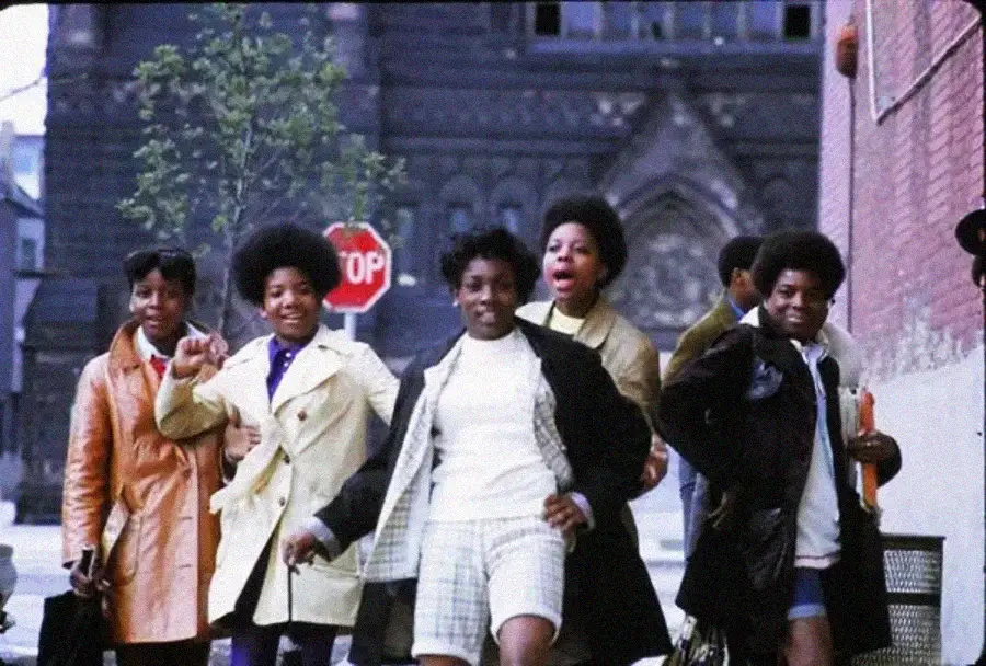 Five young people with natural hairstyles walk confidently down a city sidewalk, wearing 1970s-style coats and clothes, with a church and stop sign visible in the background.