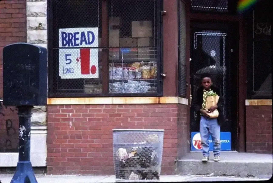 A young boy holding a paper bag stands outside a brick store with a “BREAD 5 LOAVES $1” sign in the window. A trash can and a blue mailbox are on the sidewalk in front.