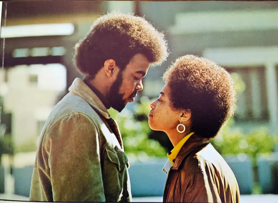 A man and woman stand face to face outdoors, gazing intently at each other. Both have natural hairstyles and wear jackets. The background is blurred, suggesting an urban setting.