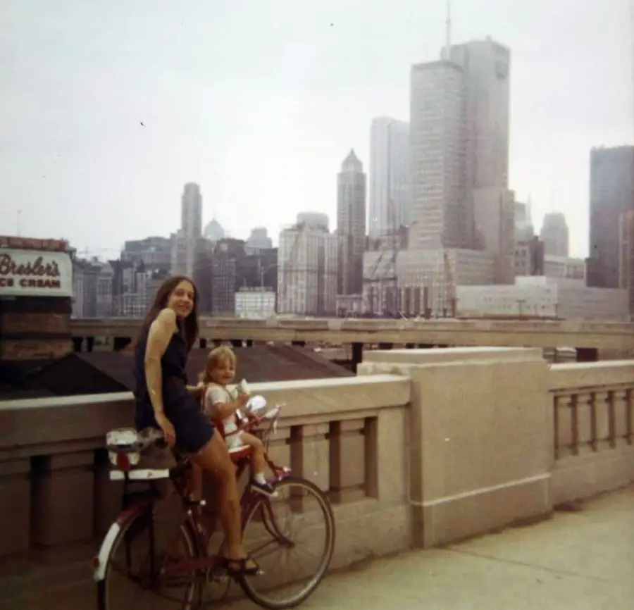 A woman and child sit on a bicycle on a city bridge with tall skyscrapers in the background on a cloudy day. A sign for Bresler’s Ice Cream is visible to the left.
