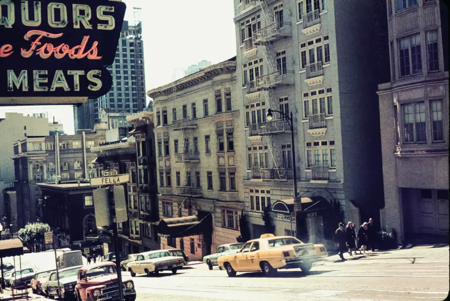 A yellow taxi drives up a steep city street lined with old buildings. A neon sign for liquors, fine foods, and meats hangs overhead. Pedestrians walk on the sidewalk.