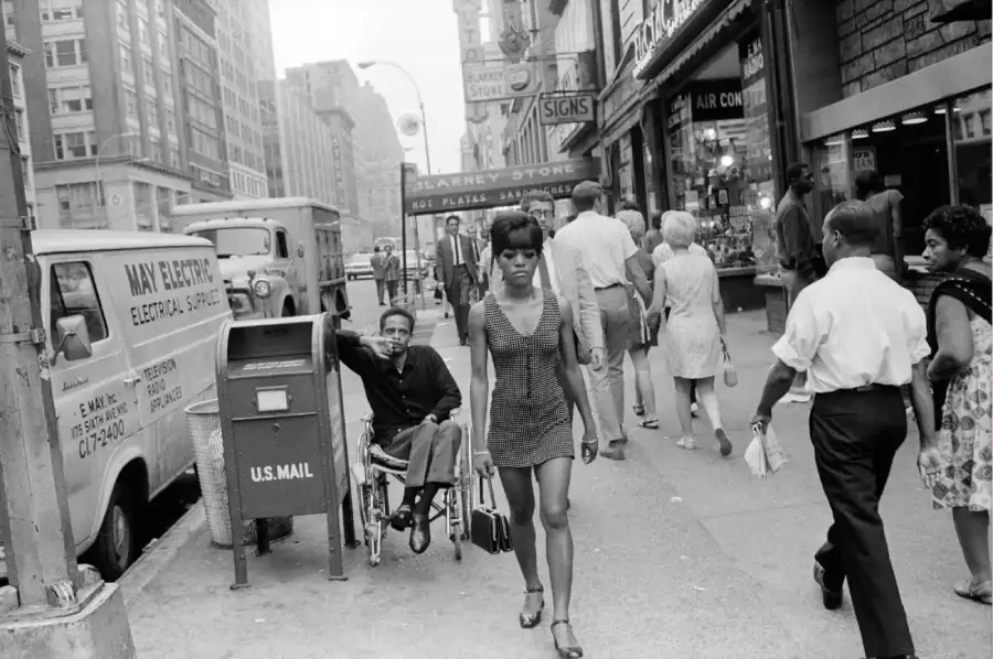 A woman in a patterned dress walks down a busy city sidewalk. People pass by storefronts, a man in a wheelchair waves, and a van and mailbox are on the street. The scene appears to be from the mid-20th century.