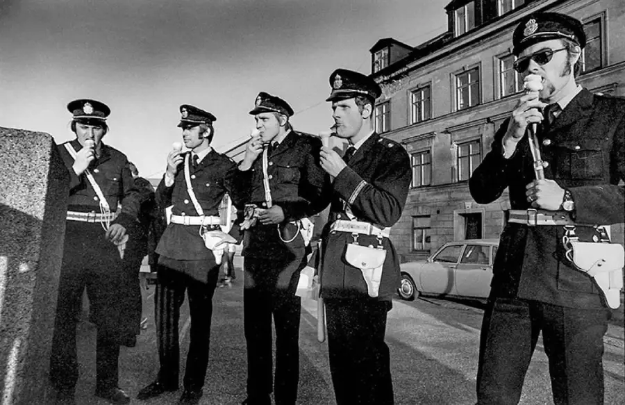 Five uniformed police officers stand outside a building, blowing into handheld breathalyzer devices. A vintage car and a large stone structure are visible in the background. The image is in black and white.
