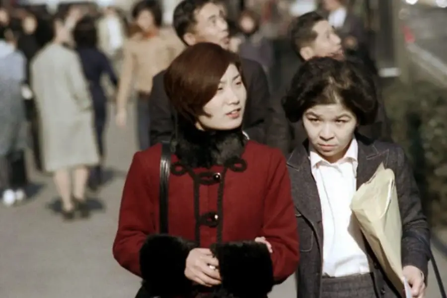 Two women walk arm in arm on a busy city sidewalk. One wears a red coat with black trim, and the other wears a suit and holds a large envelope. People and buildings are visible in the background.