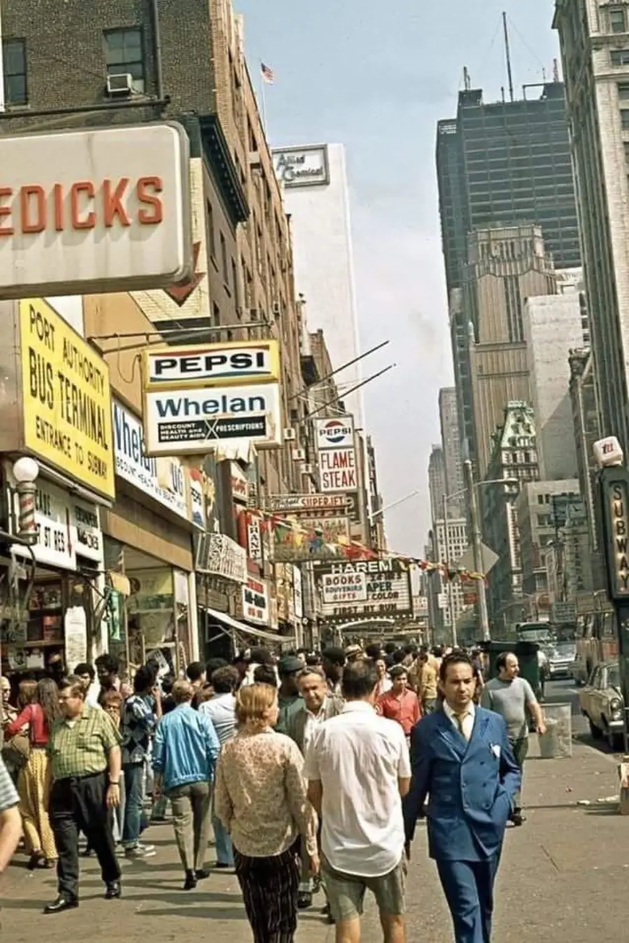 Busy city street with people walking, surrounded by tall buildings and colorful signs for shops, restaurants, and a bus terminal. Vintage cars are parked along the street. The scene captures a lively urban atmosphere.