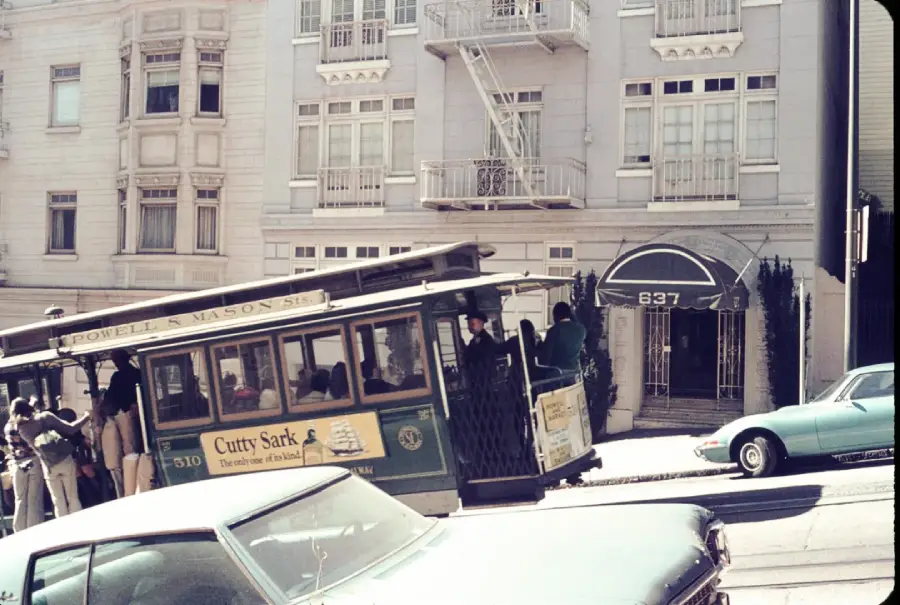 A classic San Francisco cable car travels uphill past a white building with balconies and an awning marked “837,” with parked cars visible in the foreground.