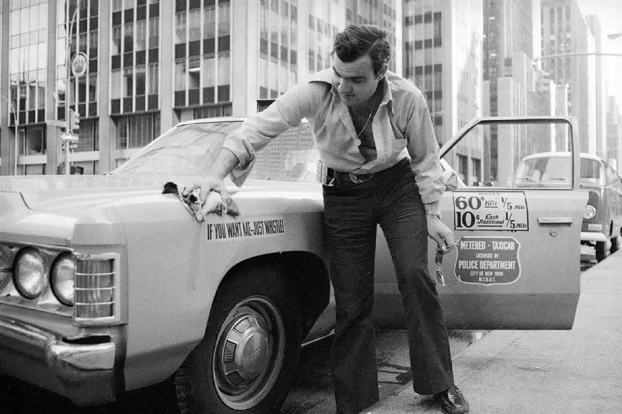 A man in a buttoned shirt leans over and wipes a yellow car labeled “Police Department” on a city street, with tall buildings and another car in the background.