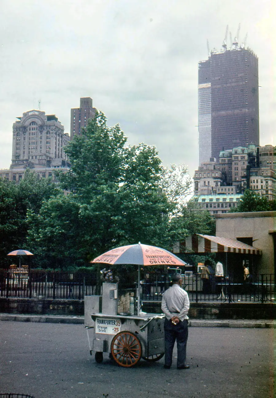A vendor stands at a hot dog cart under an umbrella in front of trees and city buildings, with a tall skyscraper under construction in the background.