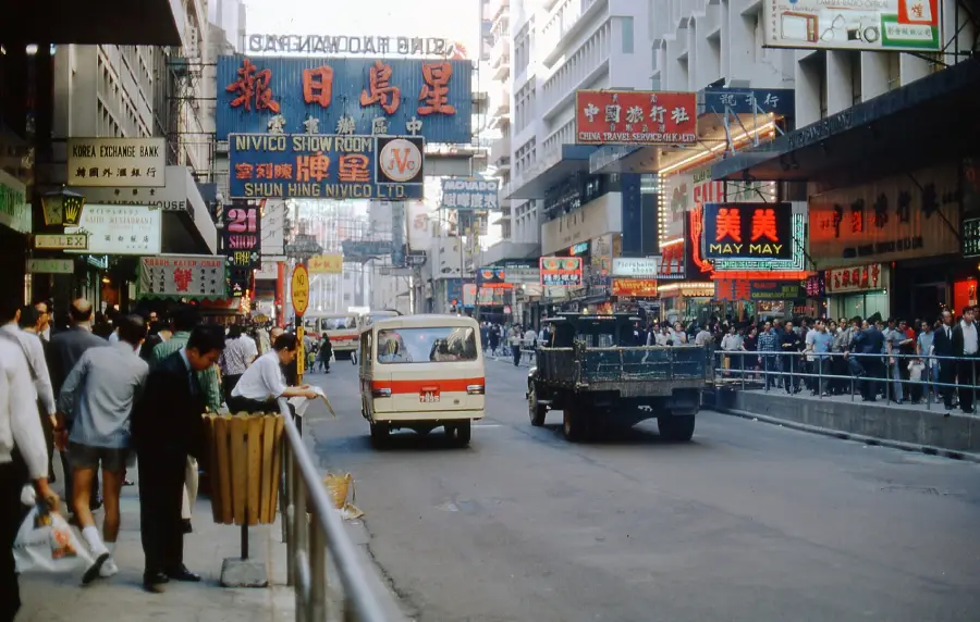 Busy city street in Hong Kong with neon signs in Chinese and English, shops, crowds of people on sidewalks, a white van, and a black truck driving down the road. Urban buildings line both sides.